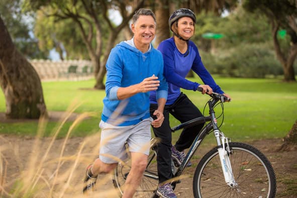 Older couple cycling together in a sunlit park, smiling — wellness in action